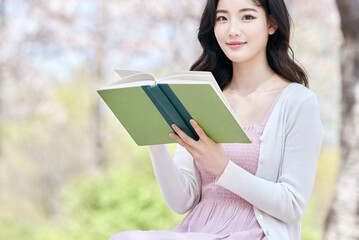Obraz premium Young Asian female reading book outdoors in spring park, wearing pink dress and white cardigan