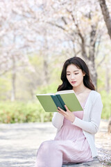 Obraz premium Young Asian female reading book outdoors in spring park, wearing pink dress and white cardigan