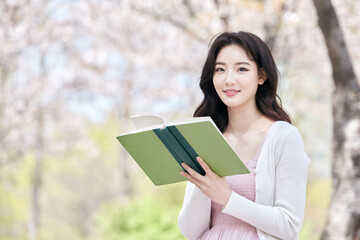 Obraz premium Young Asian female reading book outdoors in spring park, wearing pink dress and white cardigan