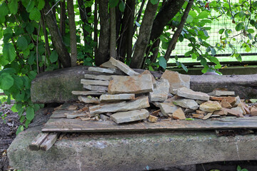 ​A pile of flat, uneven stones rests on a wooden board, which in turn lies on a massive concrete or stone base. The background consists of wooden tree trunks and lush foliage, creating a rural, constr