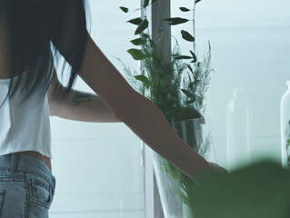Young woman placing fresh flowers into clear vase on windowsill with soft natural light creating calm mood