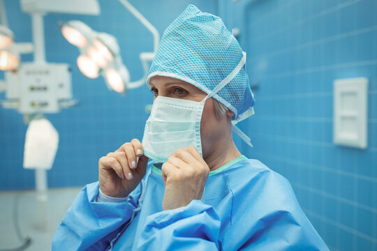 Surgeon in blue scrubs adjusting surgical mask in OR with surgical lights and control panel - Powered by Adobe