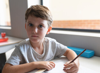 Young boy drawing in notebook at classroom table, holding pencil and showing curiosity