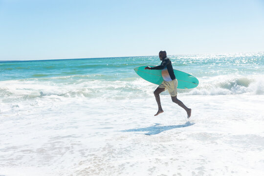 African American man carrying turquoise surfboard while walking on sunlit beach, copy space - Powered by Adobe