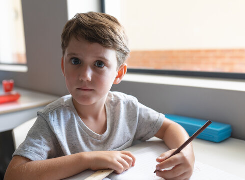 Young boy drawing in notebook at classroom table, holding pencil and showing curiosity