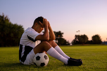 Teen soccer player sitting on grass field at dusk, holding ball under thigh near field lights