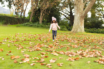 African American child running across park clearing wearing pink hat and jacket among autumn leaves