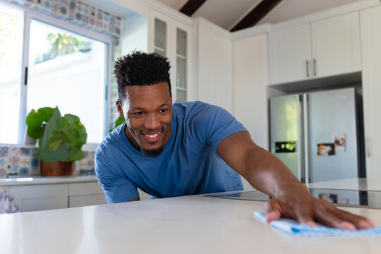 African American man leaning on home kitchen island wiping countertop with cloth under sunny window