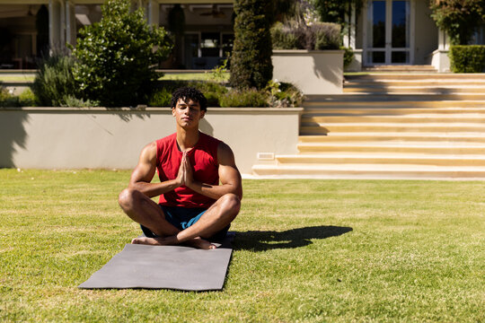 Young man meditating cross-legged on grey yoga mat in front yard, soaking up bright sunshine