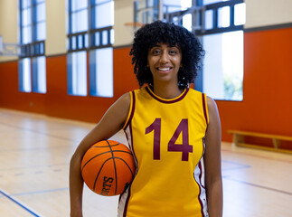 Female basketball player standing on gym court with hoop, holding basketball and wearing #14 jersey