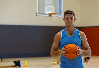 Male basketball player holding basketball on gym court, with training cones and hoop, copy space