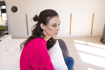 Naklejka premium Woman sitting on bed holding white pillow in bright modern bedroom, wearing pink knit sweater