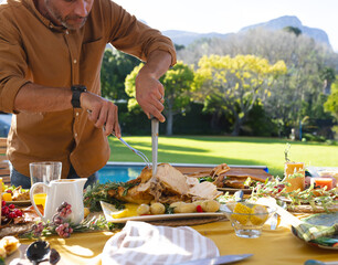 Middle-aged man carving roast turkey with knife and fork on patio table by pool, copy space