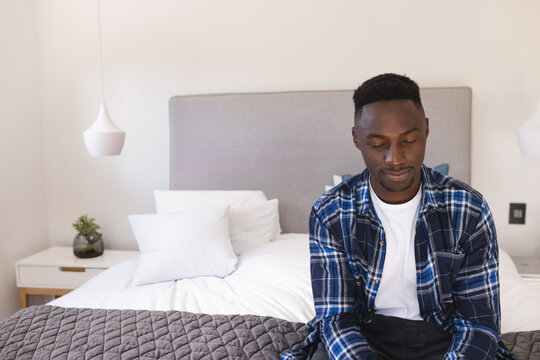 Young African American man sitting on bed edge in modern bedroom, with grey quilt, copy space