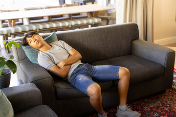 Asian man reclining on dark gray sofa in living room, with teal pillow, rug, striped bench