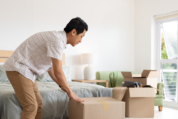 Mid adult Asian man bending over lifting cardboard box in bedroom with wooden headboard and lamp