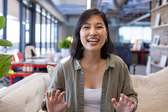 Asian woman smiling and gesturing toward camera in office lounge, with beige sofa, potted plant