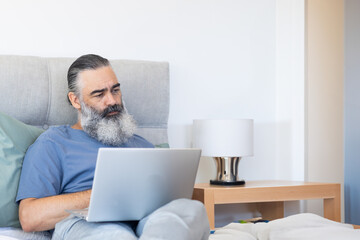 Senior man leaning against tufted headboard and working on laptop on bed, copy space