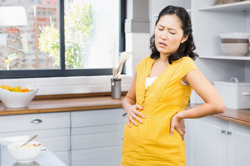 Korean pregnant woman in yellow maternity dress leaning on countertop in kitchen by fruit bowl