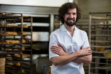 Mid adult man baker crossing arms and smiling in bakery area with metal racks of bread
