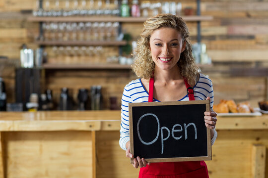 Young woman barista standing behind counter in café, holding open chalkboard sign with pastries