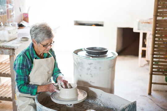 Senior female potter in apron shaping clay vessel on pottery wheel in ceramics studio, copy space