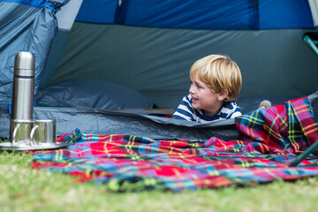 Boy child lying at blue tent entrance on plaid blanket holding metal cup and thermos