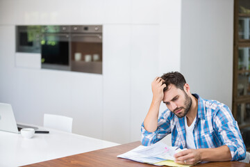 Man sitting at table in modern kitchen reviewing bills with laptop while holding head, copy space