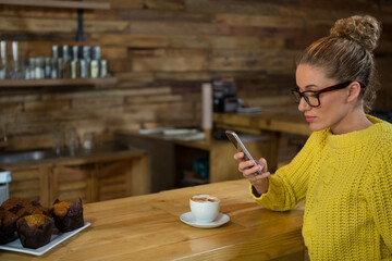 Woman wearing chunky sweater scrolling smartphone at cafe bar with latte, muffins copy space