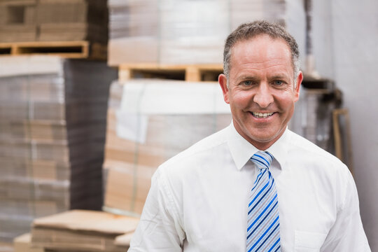 Senior man in business attire standing in warehouse among cardboard boxes and pallets, copy space - Powered by Adobe