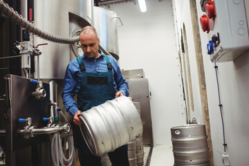 Male brewer wearing work overalls handling metal beer keg in brewery with fermentation tanks