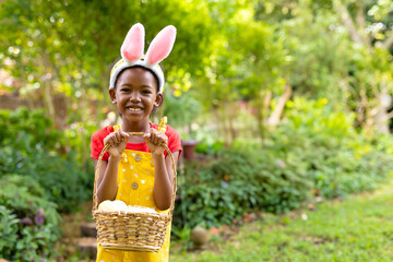 Smiling child wearing bunny ears holding basket with eggs in garden, copy space