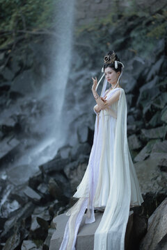A serene woman in a flowing white hanfu by a tranquil waterfall.