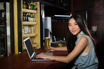 Asian woman using laptop and holding beer at bar, smiling at camera