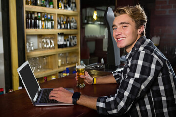 Young man using laptop and enjoying beer at bar, smiling at camera, copy space