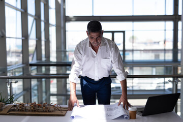 Architect reviewing blueprints at office desk with model and laptop