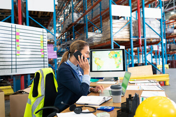 Businesswoman in warehouse talking on phone and working on computer at desk, copy space
