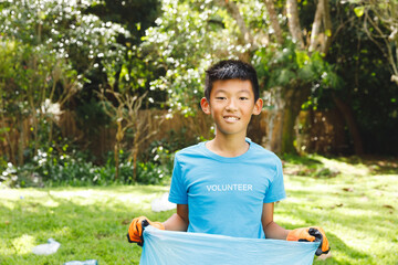 Asian boy volunteering outdoors, holding trash bag and smiling in sunny park