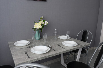 A modern dining table is set with plates, silverware, glasses, and a vase of white flowers against a gray wall
