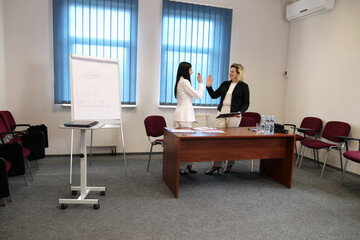 Fototapeta premium Two women in an office smile and give a high five after a successful presentation.