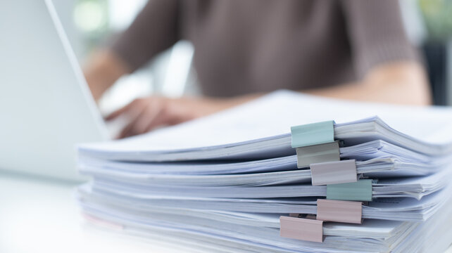 Business woman working on laptop computer with stack of paper document for searching and checking document on folder paper at busy work on desk office, close up