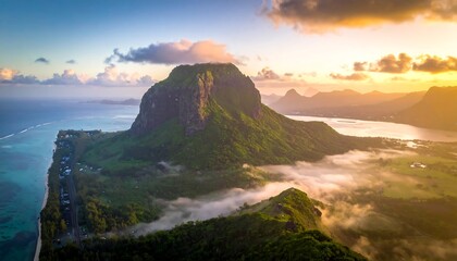 A vibrant aerial vista of a lush mountain range, bathed in the warm hues of sunrise, overlooking a tranquil valley veiled in mist and a glistening coastal expanse.