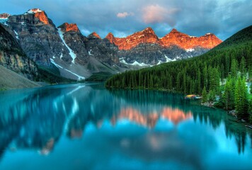 Majestic snow capped mountains reflecting in a serene turquoise lake surrounded by lush green pine forests at sunrise