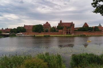 Obraz premium Historic castle overlooking tranquil river with lush greenery and cloudy sky in the background during late afternoon