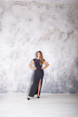 A beautiful woman standing posing in a long dark dress with a high slit on a gray studio wall background. Feminine fashion and style.