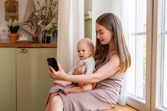 Young mother looking at gadget with baby in her arms in kitchen.
