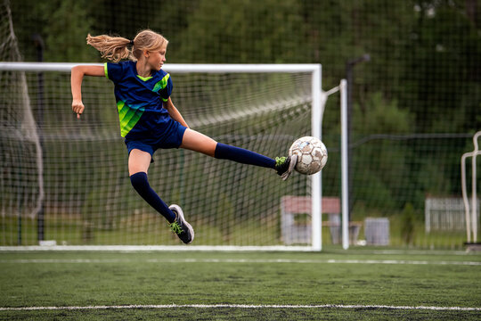 young girl playing soccer, kicking a football on a green field.