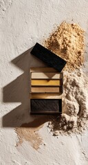 A wooden palette box, with three color blocks inside, sits atop a light surface, surrounded by piles of pale earth-toned powders.  Shadows cast by the box and powders