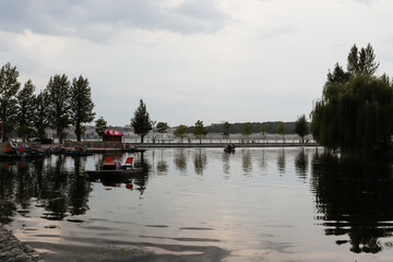 Fototapeta premium A serene lake with paddle boats and people enjoying a calm day, with the lush trees and a scenic pier reflecting on the tranquil water's surface.