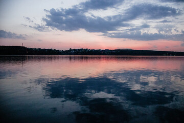 A tranquil lake reflects the soft, pastel colors of the sky at dusk, with the distant shoreline of a small village silhouetted against the horizon.